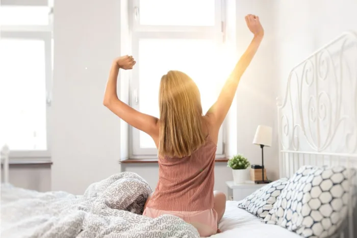 frases de bom dia: Mulher acordando e espreguiçando de frente ao uma janela ensolarada.